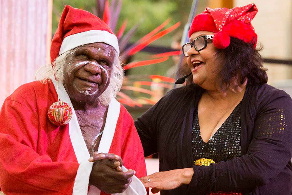 An elderly man is dressed in a red Santa outfit. The end of his cap has a striped red and green bauble attached to it. Next to him, a woman with glasses speaks into a headset and warmly grabs his arm. She is wearing a bright red cap.