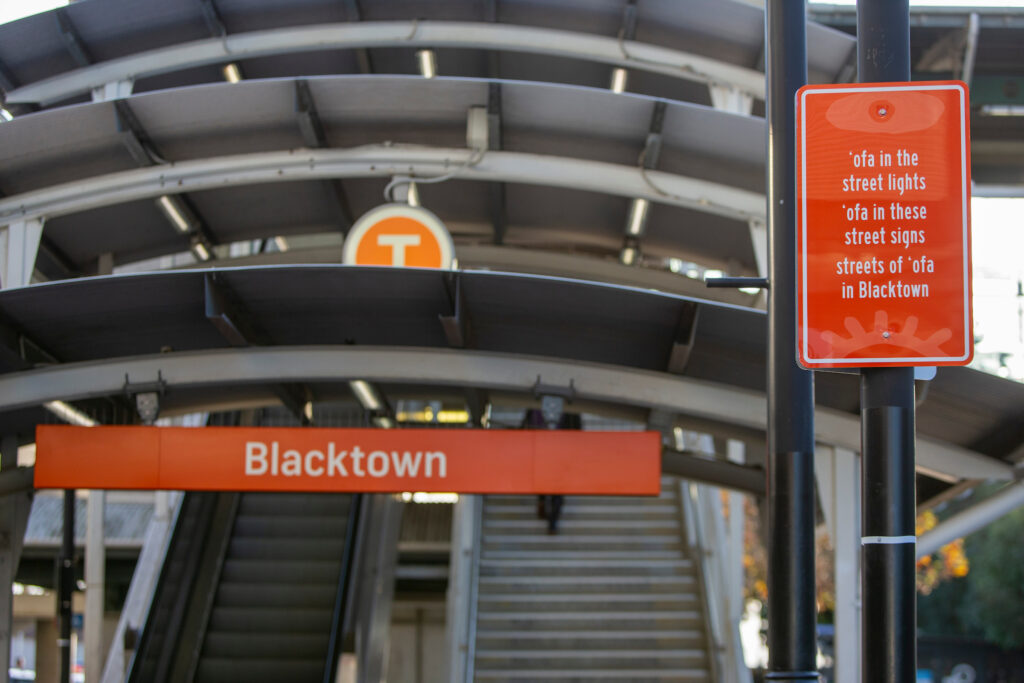 An orange street sign outside of Bankstown station with a poem. It reads " An orange street sign outside of Blacktown Station. It reads “ ‘ofa in the street lights // ‘ofa in these street signs // streets of ‘ofa in Blacktown”