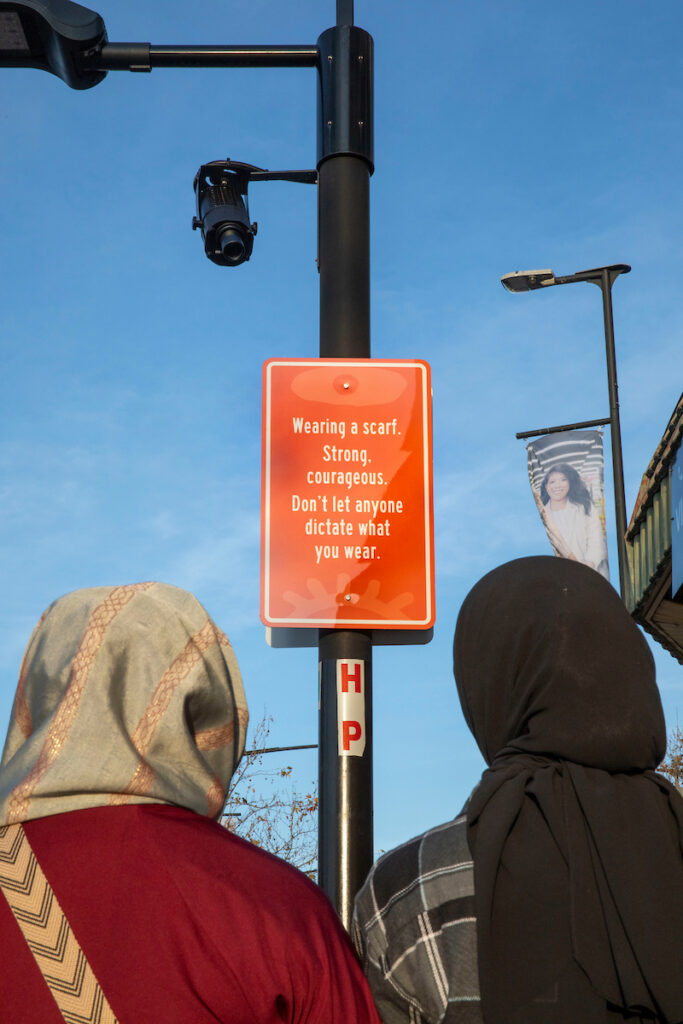 Two figures wearing a headscarf gazing at an orange street sign outside of Blacktown Station. It reads “Wearing a scarf. Strong, courageous. Don’t let anyone dictate what you wear.”