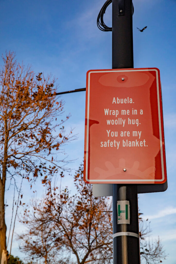 An orange street sign with a poem. It reads “Abuela. Wrap me in a woolly hug. You are my safety blanket.”