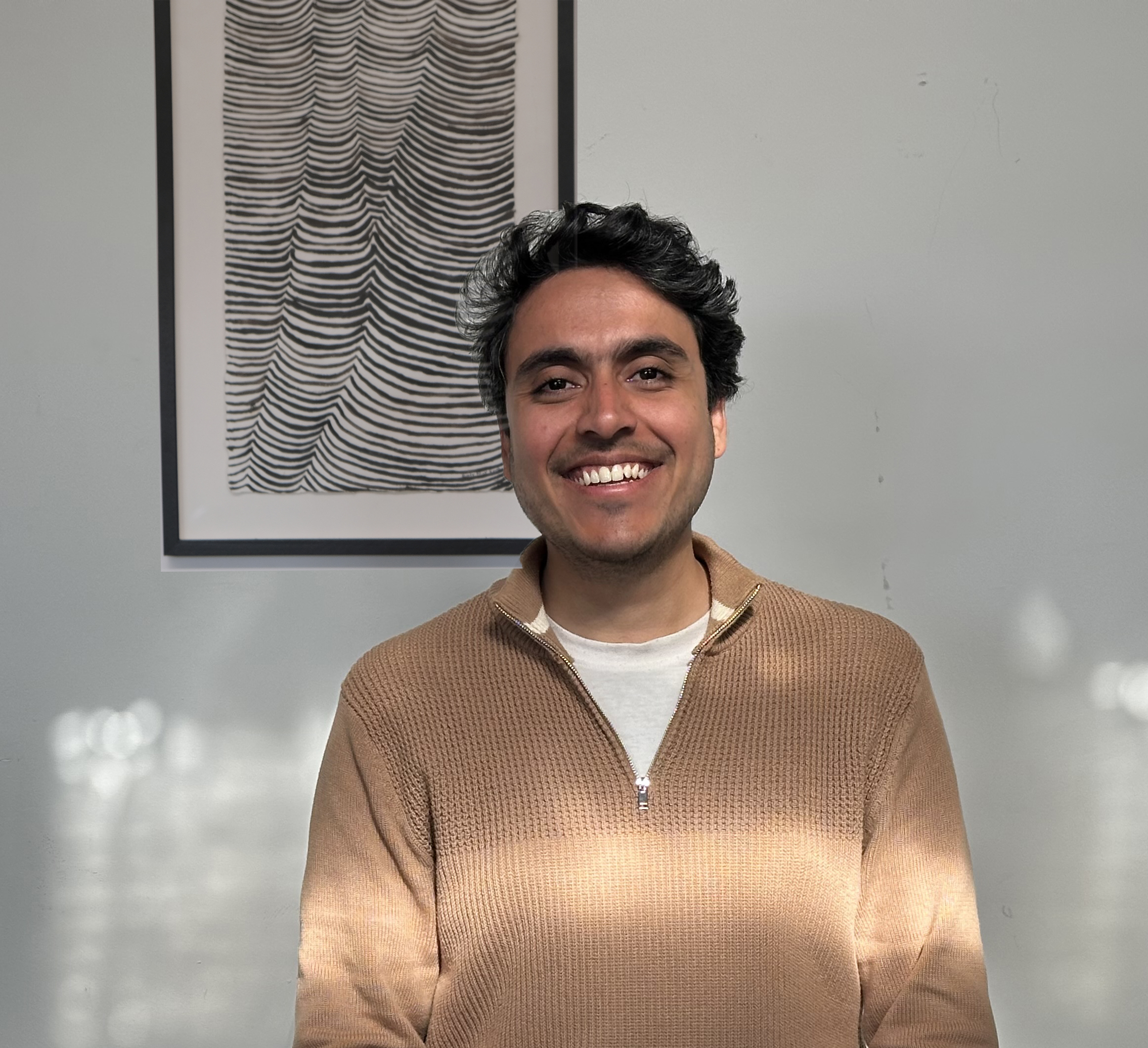 Man with a smile stands in front of a white wall with an artwork behind him. He is wearing a brown jacket and white T-shirt. There is afternoon sun on the wall and his clothes.